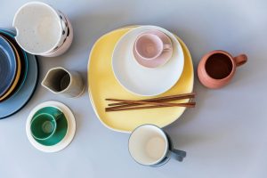 A flat lay of colorful ceramic tableware including plates, mugs, and chopsticks on a gray surface.
